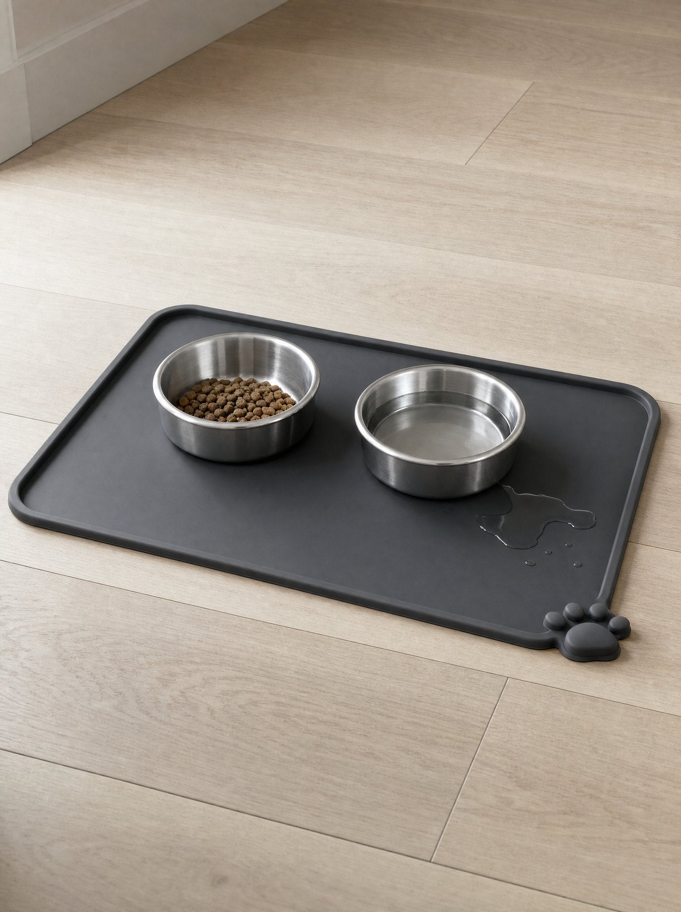 The charcoal silicone feeding mat on a wooden floor with two stainless steel bowls and a spill held on the mat